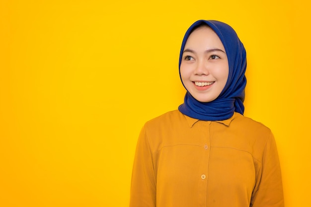 Uśmiechnięta młoda azjatycka muzułmanka ubrana na pomarańczowo, patrząca z boku na miejsce na kopię na żółtym tle