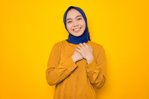 Uśmiechnięta młoda azjatycka muzułmanka ubrana na pomarańczowo, trzymająca dłonie na klatce piersiowej nad sercem na żółtym tle