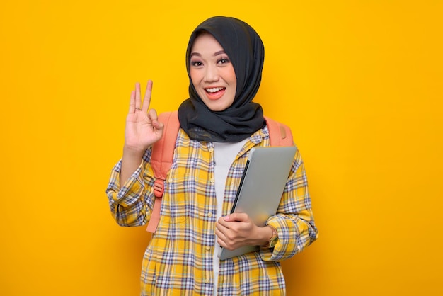 Uśmiechnięta młoda azjatycka muzułmańska studentka w kraciastej koszuli i plecaku trzymaj laptopa, wykonując zatwierdzenie ok gestem palcem na żółtym tle