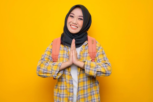 Uśmiechnięta młoda azjatycka muzułmańska studentka w zwykłych ubraniach i plecaku daje pozdrowienia ręce z dużym uśmiechem na żółtym tle Edukacja szkoła uniwersytet koncepcja kolegium