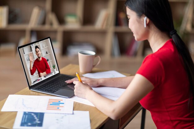 Uśmiechnięta młoda azjatycka studentka w słuchawkach ogląda laptopa z nauczycielem na ekranie, prowadzi rozmowę wideo i studiuje