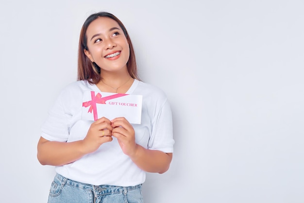 Uśmiechnięta młoda blondynka dziewczyna Azjatka ubrana w zwykły biały tshirt trzymająca kupon bonowy i patrząca na bok odizolowana na białym tle Koncepcja stylu życia ludzi