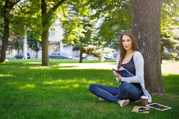 Uśmiechnięta młoda brunetka azjatyckie dziewczyny relaks na trawie i robienia notatek w parku w ciągu dnia. Picie kawy w pobliżu smartfona i tabletu. Koncepcja stylu życia i technologii