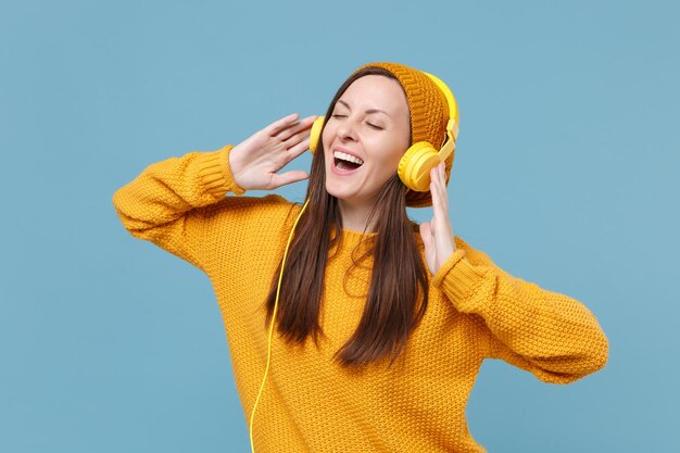 Uśmiechnięta młoda brunetka kobieta dziewczyna w kapeluszu żółty sweter pozowanie na białym tle na niebieskim tle portret studio. Koncepcja życia szczere emocje ludzi. Makieta miejsca na kopię. Słuchaj muzyki przez słuchawki.