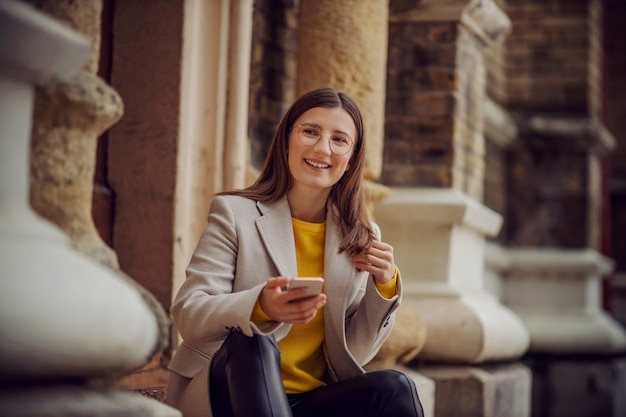 Uśmiechnięta młoda brunetka w żółtym swetrze siedzi na schodach w centrum miasta i czeka na wysłanie wiadomości