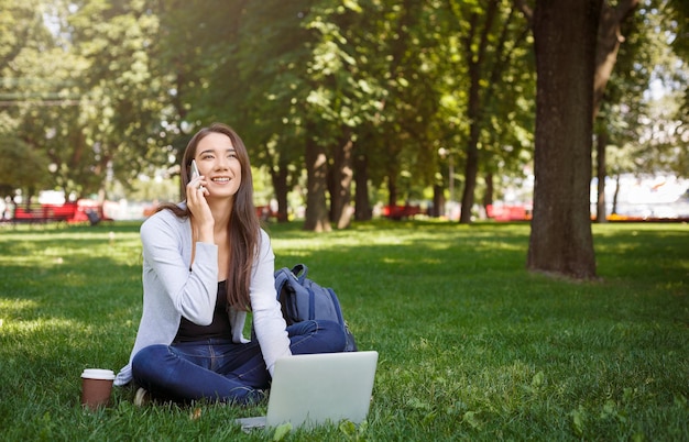 Uśmiechnięta młoda brunetki kobieta na zielonej trawie. Praca z laptopem i rozmowa przez telefon w parku w ciągu dnia. Praca jako freelancer w wolnym czasie. Koncepcja stylu życia i technologii