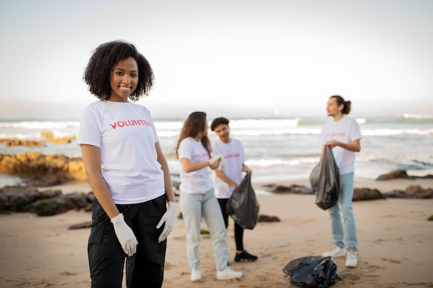 Uśmiechnięta młoda czarna dama i różni przyjaciele wolontariusze w rękawiczkach z koszulkami i workami na śmieci sprzątają