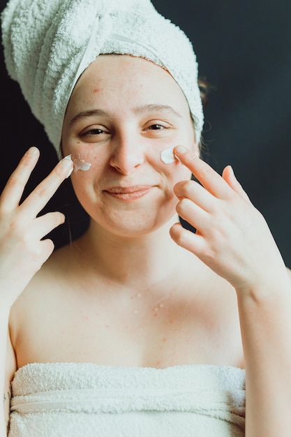 Uśmiechnięta młoda dorosła kobieta z ręcznikiem używająca kremu do twarzy po prysznicu Pielęgnacja skóry odnowy biologicznej radosnej twarzy, blasku skóry i kremu przeciwsłonecznego Codzienna rutyna chroniąca delikatną skórę