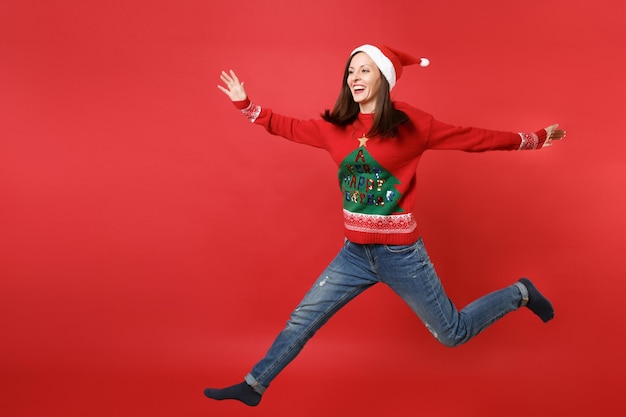 Uśmiechnięta młoda dziewczyna Santa w sweter z dzianiny Boże Narodzenie kapelusz skoki rozłożone ręce i nogi na białym tle na jasnym czerwonym tle. Szczęśliwego nowego roku 2019 celebracja party wakacje koncepcja. Makieta miejsca na kopię.