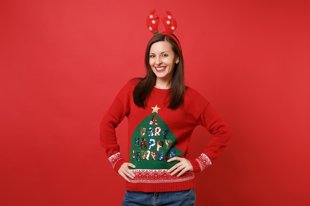 Uśmiechnięta młoda dziewczyna Santa w zabawne ozdobne rogi jelenia na głowie stojącej z ramionami akimbo na białym tle na jasnym czerwonym tle. Szczęśliwego nowego roku 2019 celebracja party wakacje koncepcja. Makieta miejsca na kopię.
