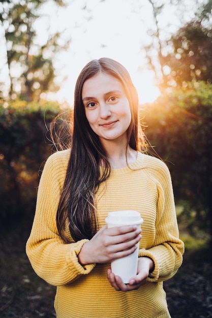Uśmiechnięta młoda dziewczyna ubrana w żółty sweter i trzymająca jednorazową filiżankę kawy o zachodzie słońca w parku