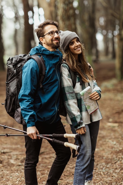 Uśmiechnięta młoda europejska para turystów w kurtkach z plecakiem, kijami trekkingowymi i spacerem po mapie