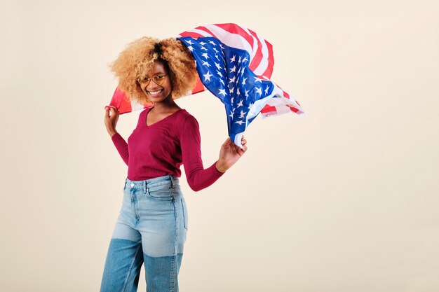 Uśmiechnięta młoda kobieta afro machająca flagą USA, stojąc na na białym tle. Dzień Niepodległości USA i koncepcja patriotyzmu.