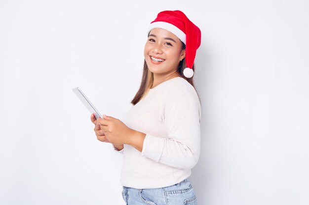 Uśmiechnięta młoda kobieta Azji w kapeluszu Boże Narodzenie przy użyciu cyfrowego tabletu izolowanych na białym tle Indonezyjczycy świętują koncepcję Bożego Narodzenia