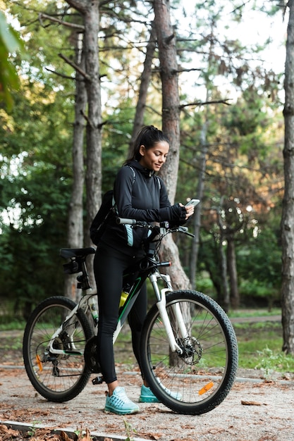 Uśmiechnięta młoda kobieta, jazda na rowerze w parku, telefon komórkowy