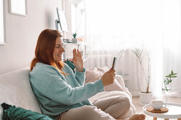Uśmiechnięta młoda kobieta korzystająca z telefonu komórkowego, siedząc na kanapie w domu