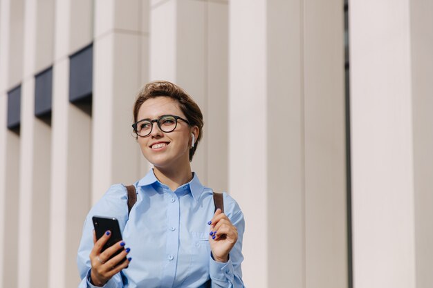 Uśmiechnięta młoda kobieta na sobie niebieską koszulę i okulary o czat wideo na smartfonie. Szczęśliwa kobieta stojąca na zewnątrz i za pomocą nowoczesnego gadżetu.