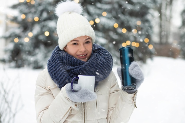 Uśmiechnięta młoda kobieta pije kawę z termosu w winter park podczas opadów śniegu. Miejsce na tekst