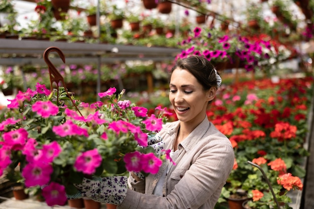 Uśmiechnięta młoda kobieta pracująca w ogrodzie kwiatowym, trzymająca doniczki i ciesząca się pięknymi i kolorowymi kwiatami.