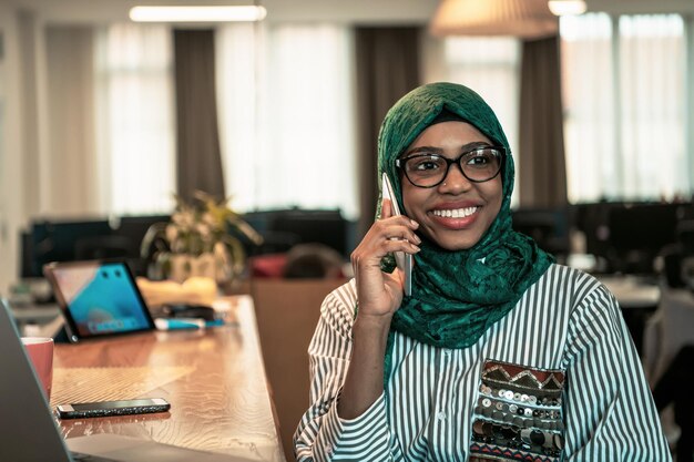 Uśmiechnięta Młoda Kobieta Rozmawia Na Smartfonie Działa Na Laptopie W Myśleniu Na Odległość. Klient Zadowolony African American Przy Użyciu Nowoczesnych Gadżetów Telefon I Komputer W Domowym Biurze. Wysokiej Jakości Zdjęcia