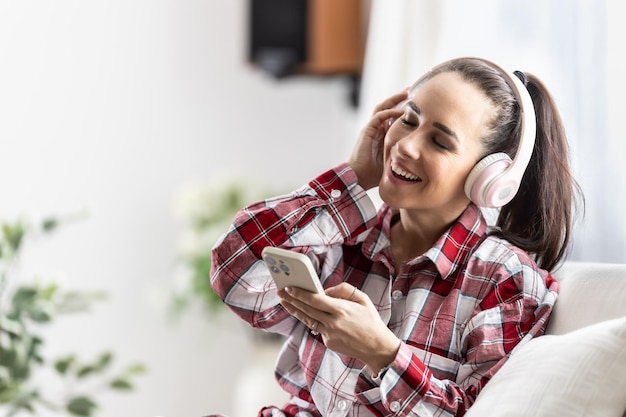 Uśmiechnięta młoda kobieta siedzi na kanapie z telefonem komórkowym i słucha muzyki w słuchawkach