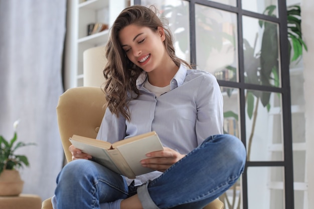Uśmiechnięta młoda kobieta siedzi w fotelu w salonie i czyta książkę.