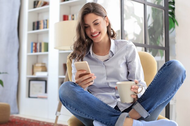 Uśmiechnięta młoda kobieta siedzi w fotelu w salonie i za pomocą telefonu komórkowego.