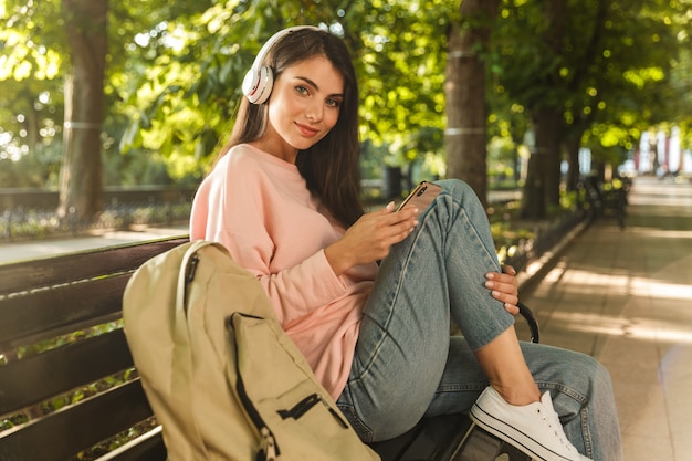 Uśmiechnięta młoda kobieta słucha muzyki przez słuchawki, siedząc na ławce w parku miejskim i trzymając telefon komórkowy