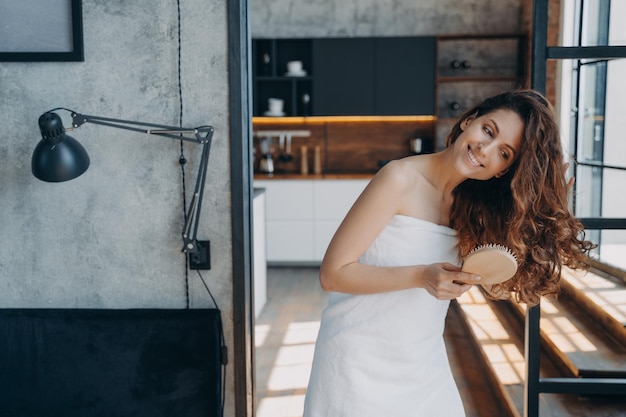Uśmiechnięta młoda kobieta szczotkująca zdrowe długie brązowe włosy po prysznicu w domu Zabieg pielęgnacyjny włosów