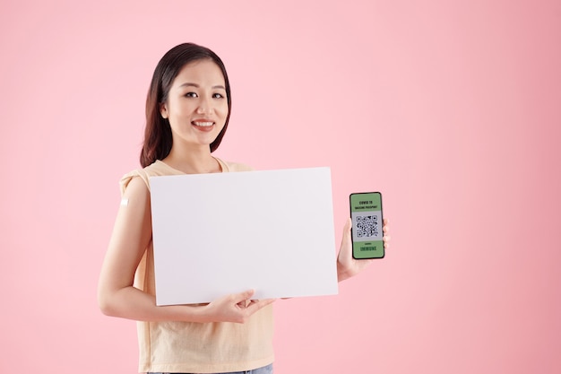 Uśmiechnięta młoda kobieta trzymająca białą tabliczkę i smartfona z kodem qr potwierdzającym, że została zaszczepiona