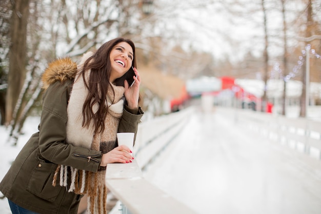 Uśmiechnięta młoda kobieta używa telefon w parku przy zimnym zima dniem