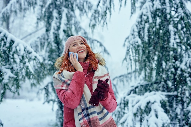 Uśmiechnięta młoda kobieta używa telefon w parku przy zimnym zima dniem