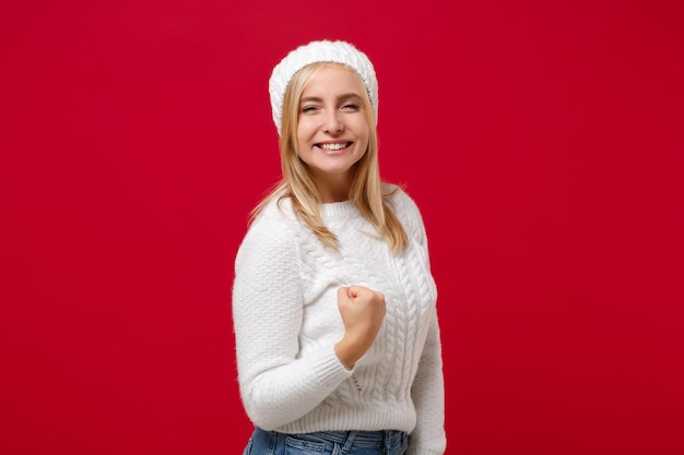 Zdjęcie uśmiechnięta młoda kobieta w białym pulowerze kapeluszu odizolowywającym na czerwonym tle pracowniany portret. zdrowy styl życia ludzi szczere emocje, koncepcja zimnej pory roku. makieta miejsca na kopię. robiąc gest zwycięzcy.