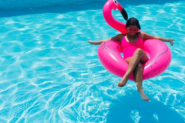 Uśmiechnięta młoda kobieta w bikini relaksuje się na nadmuchiwanym różowym flamingu w basenie. Atrakcyjna kobieta w stroju kąpielowym leży w słońcu na tropikalnych wakacjach. Kobieta opalająca się w ośrodku.