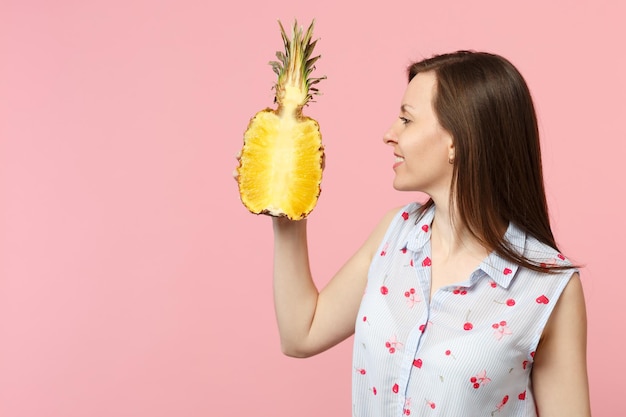 Uśmiechnięta młoda kobieta w letnie ubrania trzymaj w ręku, patrząc na połowę świeżych dojrzałych owoców ananasa na białym tle na różowym pastelowym tle. Ludzie żywy styl życia zrelaksować koncepcję wakacji. Makieta miejsca na kopię.