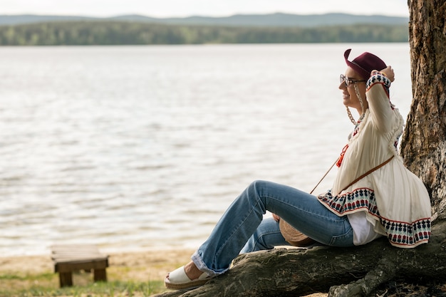 Uśmiechnięta młoda kobieta w okularach i bluzce hippie siedzi na drzewie i trzymając kapelusz, ciesząc się wiatrem w pobliżu jeziora