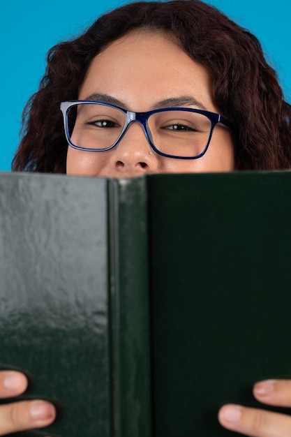 Uśmiechnięta młoda kobieta w okularach ukrywa połowę twarzy za książką w twardej oprawie