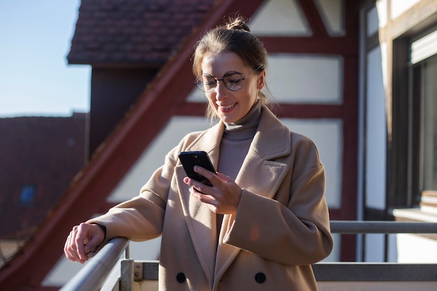 Uśmiechnięta młoda kobieta w płaszczu patrząc na telefon stojąc na tarasie