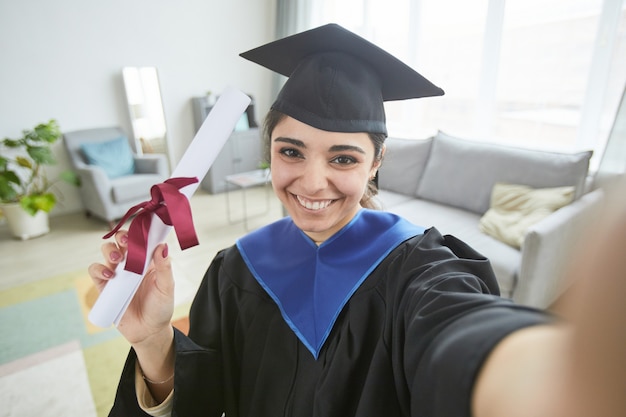 Uśmiechnięta młoda kobieta w sukni ukończenia szkoły podczas robienia selfie w domu lub na blogu wideo