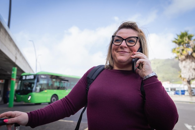 Uśmiechnięta młoda kobieta wykonuje telefon, czekając na autobus na dworcu