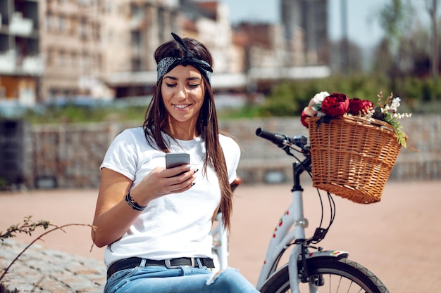 Uśmiechnięta młoda kobieta wysyła SMS-y na smartfonie na ulicy miasta, w słoneczny dzień, obok roweru z koszem kwiatów.