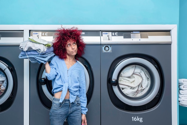 Uśmiechnięta młoda kobieta z czerwonymi włosami afro trzymająca ubrania w niebieskiej automatycznej pralni