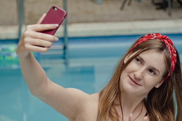 Uśmiechnięta młoda kobieta z telefonem na bryczce. Relaksujący dzień w spa przy basenie latem robiący sobie zdjęcie lub wideorozmowy z chłopakiem