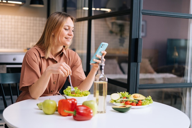 Uśmiechnięta Młoda Kobieta Za Pomocą Telefonu Komórkowego Podczas Jedzenia Sałatki W Przytulnej Drewnianej Kuchni. Pojęcie Zdrowego Odżywiania.