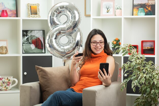 Uśmiechnięta młoda ładna dziewczyna w okularach optycznych trzymająca kieliszek wina i patrząca na telefon siedzący na fotelu w salonie w marcowy międzynarodowy dzień kobiet