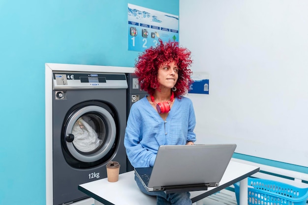 Uśmiechnięta młoda Latynoska z rudymi włosami afro siedzi przy stole w niebieskiej pralni, pracując na swoim laptopie, czekając na zrobienie prania, z czerwonymi słuchawkami na szyi