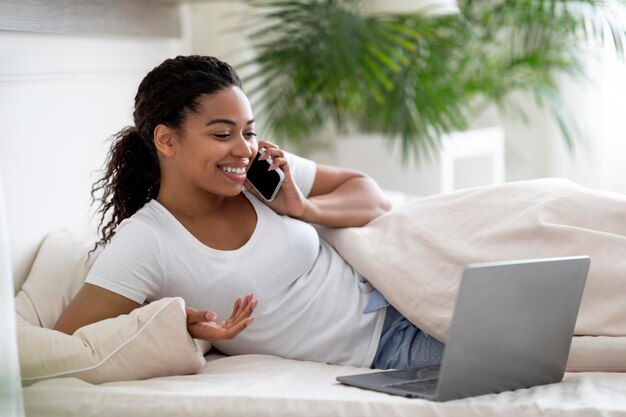 Uśmiechnięta młoda murzynka relaksuje w łóżku z telefonem komórkowym i laptopem