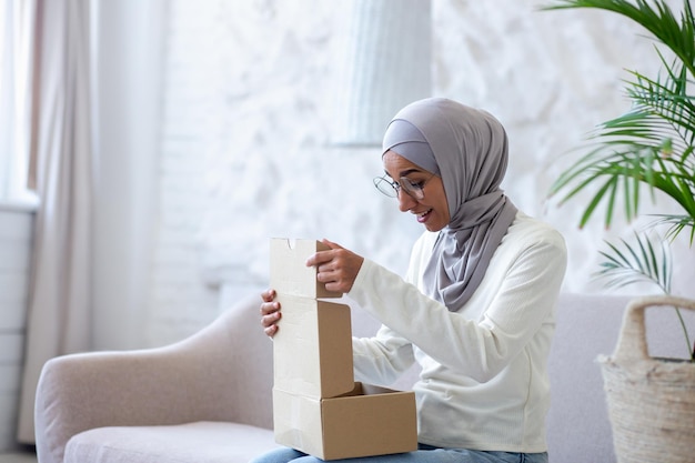 Uśmiechnięta młoda muzułmanka w hidżabie siedzi na kanapie i trzyma paczkę otwierającą pudełko, z której jest zadowolona