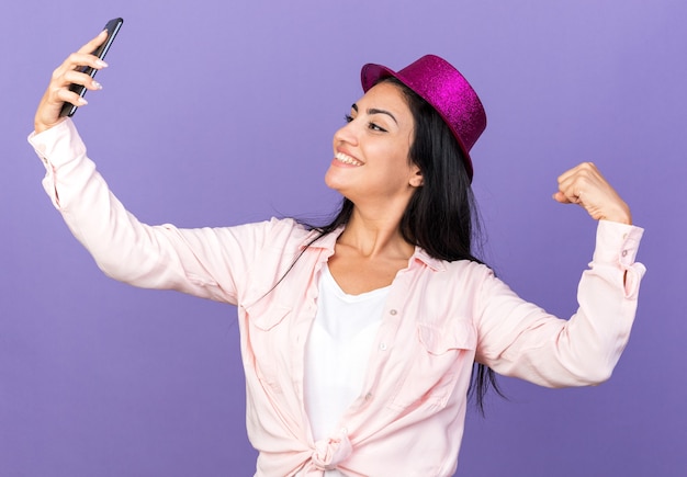 Uśmiechnięta Młoda Piękna Dziewczyna W Imprezowym Kapeluszu Robi Selfie Pokazując Gest Tak Na Niebieskiej ścianie