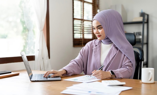 Uśmiechnięta muzułmańska bizneswoman używająca laptopa analizującego wykres biznesowy siedząca przy biurku w nowoczesnej przestrzeni biurowej